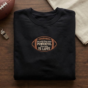 May include: Black sweatshirt with a crew neck and an embroidered football design. The football graphic has the text "the only thing more POWERFUL than hate IS LOVE" in a brown and tan color scheme. A football and whistle are visible in the background.
