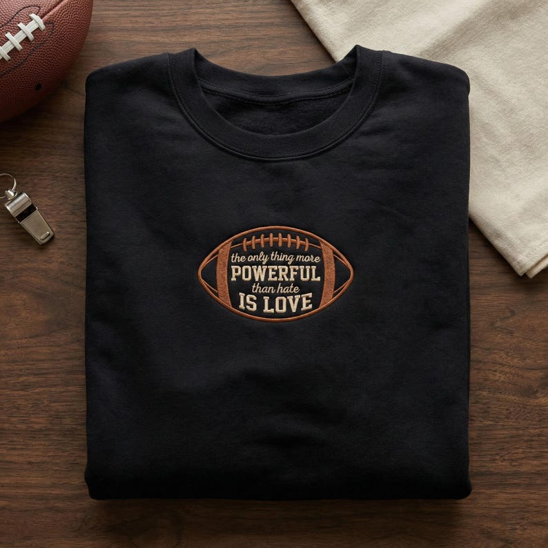 May include: Black sweatshirt with a crew neck and an embroidered football design. The football graphic has the text "the only thing more POWERFUL than hate IS LOVE" in a brown and tan color scheme. A football and whistle are visible in the background.