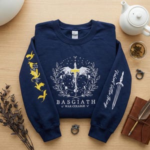 May include: Navy blue sweatshirt with white and yellow graphic design. The design includes a sword, wings, and the text "BASGIATH WAR COLLEGE". The sleeves feature additional text and illustrations of dragons and a sword.