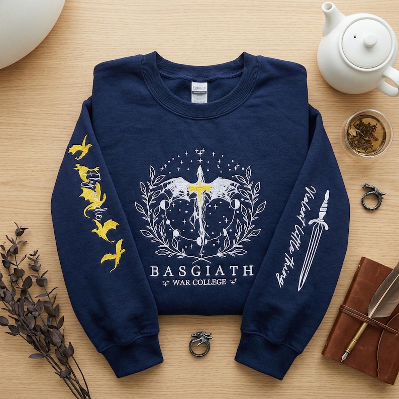 May include: Navy blue sweatshirt with white and yellow graphic design. The design includes a sword, wings, and the text "BASGIATH WAR COLLEGE". The sleeves feature additional text and illustrations of dragons and a sword.