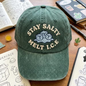 May include: A green baseball cap with the embroidered phrase "STAY SALTY MELT I.C.E." above an ice cube design. The hat is made of a textured fabric and has a curved brim. The cap is sitting on a wooden surface.