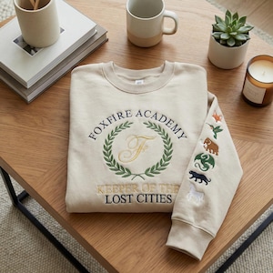 May include: Beige sweatshirt with embroidered text and graphics. The front features "FOXFIRE ACADEMY" above a laurel wreath with a gold "F" and "KEEPER OF THE LOST CITIES" below. The right sleeve has animal and plant graphics.