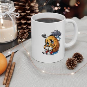 Peut inclure: Un mug en céramique blanche orné d'un dessin représentant une pomme de terre triste qui pleure sous un petit nuage de pluie gris. La pomme de terre porte des gants blancs et des baskets rouges et blanches, avec des larmes bleues au sol.