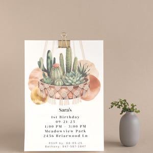Pode incluir: Um convite impresso para o 1º aniversário de Sara, com uma ilustração em aquarela de cactos em um vaso de macramê. O convite inclui detalhes do evento e informações de RSVP, preso por um clip dourado.