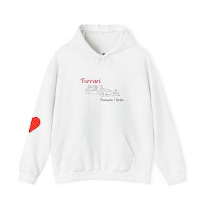 Może przedstawiać: Biała bluza z kapturem z przednią kieszenią i kapturem ze sznurkiem. Bluza z kapturem ma napis "Ferrari" w czerwonym skrypcie nad rysunkiem linii samochodu wyścigowego. Czerwone serce znajduje się na lewym rękawie. Tekst "Formula 1 Girlie" znajduje się poniżej.