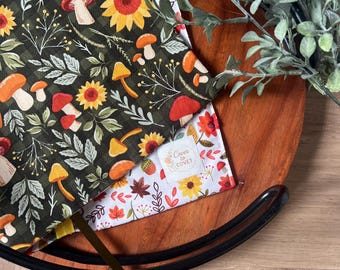 May include: Two fabric items with autumn-themed patterns. One features mushrooms, sunflowers, and leaves on a dark green background. The other has a floral design with the text "Cover to Covet". The items are on a wooden tray with a black rim, with greenery.