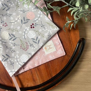 May include: Two fabric book covers with floral patterns. One cover is gray with white, pink, and green floral designs. The other is pink with white floral designs. The text "Cover to Covet" is on a small label on the pink cover.