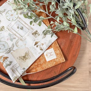 May include: Two decorative fabric napkins with botanical and vintage-inspired illustrations, including mushrooms, butterflies, and framed artwork. One napkin is white with a detailed print, and the other is a solid orange color. The napkins are displayed on a wooden tray.