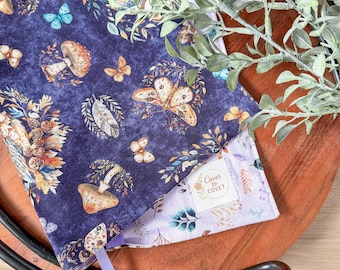 May include: A book sleeve with a dark blue floral pattern featuring butterflies, mushrooms, and foliage. The interior is a light purple with a floral design. A purple ribbon bookmark is visible. The product is labeled "Cover to Covet."