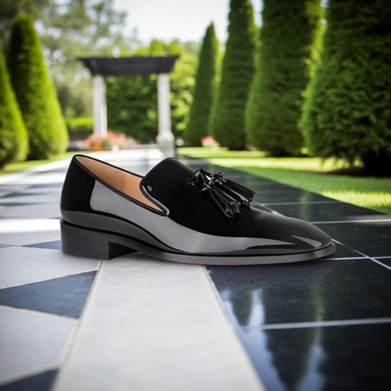 May include: A black patent leather loafer with a small heel and tassel detail. The shoe has a glossy finish and a classic design, suitable for formal or semi-formal occasions. The shoe is displayed on a black and white tiled surface.