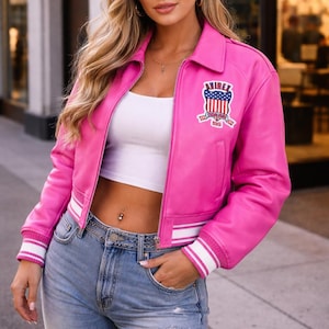 May include: A bright pink leather jacket with a white collar and cuffs. The jacket has a zipper closure and a patch on the chest with the text "AIREX" and a patriotic design. The jacket is worn with a white top and blue jeans.