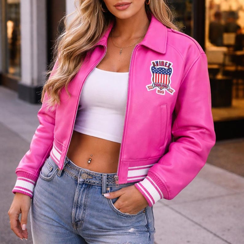 May include: A bright pink leather jacket with a white collar and cuffs. The jacket has a zipper closure and a patch on the chest with the text "AIREX" and a patriotic design. The jacket is worn with a white top and blue jeans.
