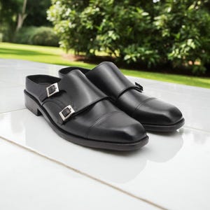 May include: A pair of black leather mule shoes with double monk straps and silver buckles. The shoes have a low heel and a rounded toe. The background is a blurred view of green foliage and a white surface.
