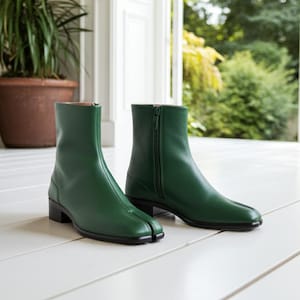 May include: A pair of green leather ankle boots with a split toe design. The boots have a side zipper and a small, black heel. The boots are displayed on a white surface.