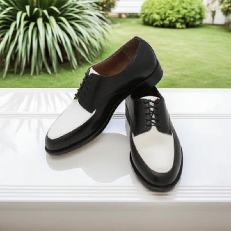 May include: A pair of two-tone dress shoes. The shoes are black with white toe caps and laces. The shoes are displayed on a white surface with a green background.