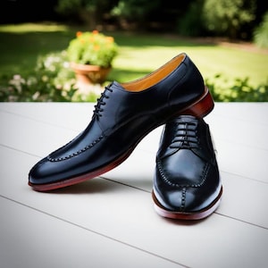 Puede incluir: Un par de zapatos de vestir de cuero negro con un dise&ntilde;o cl&aacute;sico. Los zapatos tienen cierre de cordones, punta redonda y suela roja. Se exhiben sobre una superficie blanca con un fondo verde borroso.