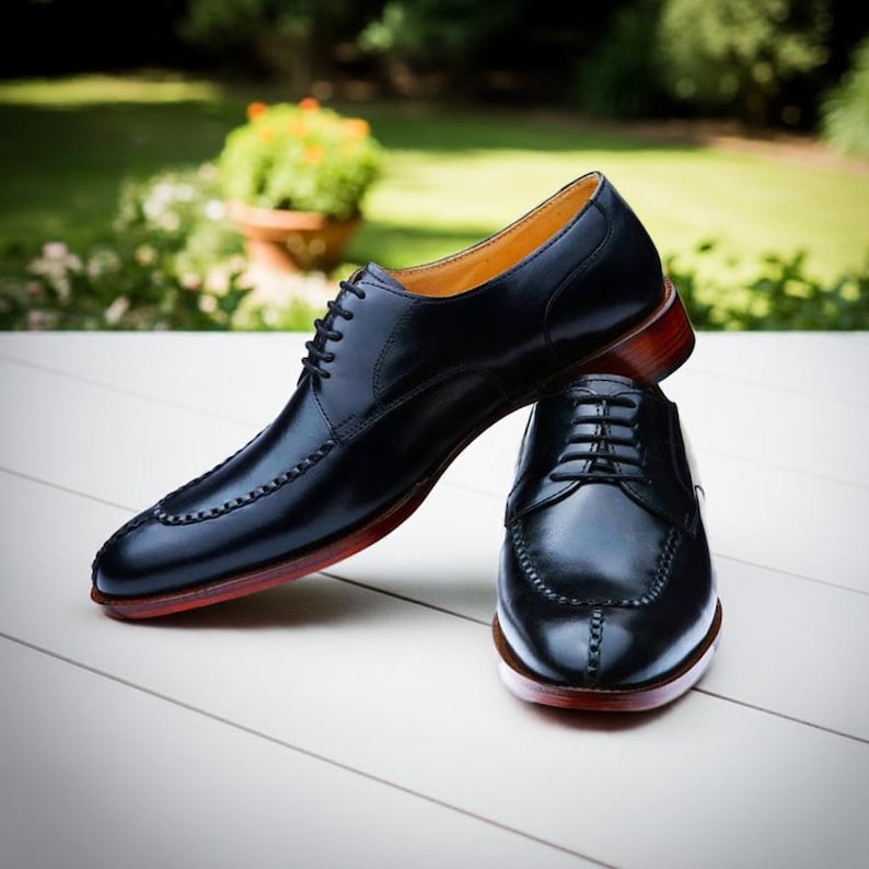 Puede incluir: Un par de zapatos de vestir de cuero negro con un dise&ntilde;o cl&aacute;sico. Los zapatos tienen cierre de cordones, punta redonda y suela roja. Se exhiben sobre una superficie blanca con un fondo verde borroso.