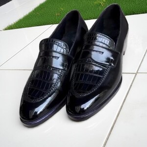 Puede incluir: Un par de mocasines de cuero negro brillante con un patr&oacute;n texturizado, similar al de cocodrilo, en la pala y la correa. Los zapatos tienen un dise&ntilde;o cl&aacute;sico de mocas&iacute;n y un acabado pulido, adecuados para vestimenta formal o de negocios.