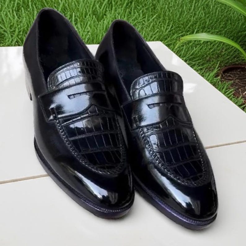 Puede incluir: Un par de mocasines de cuero negro con un acabado brillante. Los zapatos presentan un patr&oacute;n texturizado, similar al de un cocodrilo, en la pala y un dise&ntilde;o cl&aacute;sico de mocas&iacute;n. Los zapatos se exhiben sobre una superficie blanca con un fondo verde.