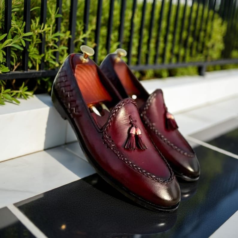 May include: A pair of burgundy leather loafers with braided trim and tassel accents. The shoes have a classic design with a polished finish, showcasing a rich color and elegant details. The loafers are displayed on a black and white checkered surface.