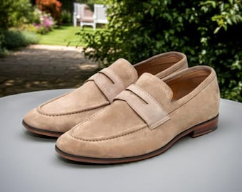 May include: A pair of tan suede loafers with a classic design. The shoes feature a rounded toe, a strap across the top, and a brown sole. The loafers are displayed on a light gray surface, with a blurred outdoor background.