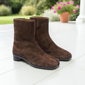 Puede incluir: Un par de botines de ante marrón con diseño de puntera dividida. Los botines tienen un tacón bajo y cierre de cremallera. Los botines se exhiben sobre una superficie de baldosas blancas, con un fondo borroso de vegetación y flores.