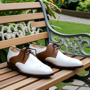 Può includere: Un paio di scarpe eleganti in pelle bicolore marrone e bianca, con lacci neri e un classico design a coda di rondine. Le scarpe sono esposte su una panchina di legno con una cornice metallica decorativa, mostrando uno stile senza tempo.