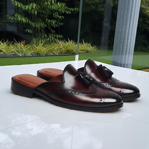 Könnte beinhalten: Ein Paar bordeauxfarbene Leder-Slipper mit Quasten-Detail. Die Schuhe haben eine braune Lederinnensohle und eine schwarze Sohle. Auf einer weißen Oberfläche mit verschwommenem Hintergrund aus Grün.