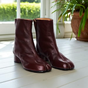 Op de afbeelding: Een paar bordeauxrode leren enkellaarsjes met een split-teen ontwerp. De laarzen hebben een lage blokhak en een ritssluiting aan de zijkant. De binnenvoering is lichtbruin. De laarzen staan op een witte ondergrond.