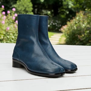 May include: A pair of blue leather ankle boots with a split-toe design. The boots have a low, black heel and a smooth, textured finish. The boots are displayed on a white surface with a blurred green background.