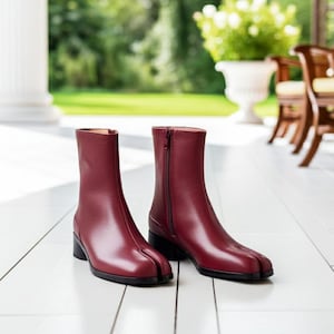Könnte beinhalten: Ein Paar burgunderfarbene Leder-Ankle Boots mit geteiltem Zehendesign. Die Stiefel haben einen seitlichen Reißverschluss, einen kleinen Blockabsatz und ein glattes Finish. Die Stiefel werden auf einer weißen Oberfläche präsentiert.