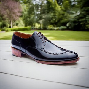 Puede incluir: Un zapato de vestir de cuero negro brillante. El zapato presenta cordones marrones, un dise&ntilde;o cosido decorativo en la puntera y un tac&oacute;n de madera. El fondo es borroso y verde.