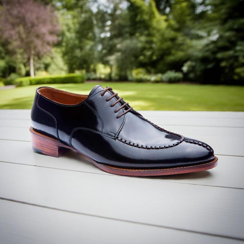 Puede incluir: Un zapato de vestir de cuero negro brillante. El zapato presenta cordones marrones, un dise&ntilde;o cosido decorativo en la puntera y un tac&oacute;n de madera. El fondo es borroso y verde.