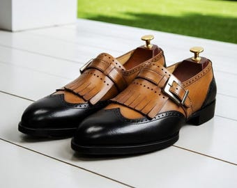 May include: A pair of two-tone leather dress shoes. The shoes feature a black toe and heel, with a tan upper and fringe detail. Each shoe has a buckle closure and is set on a white surface with a green background.
