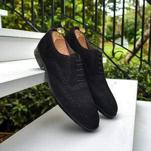 May include: A pair of black suede dress shoes with intricate brogue detailing. The shoes have a classic Oxford design with a lace-up closure and are displayed on a white surface. The image showcases the shoes' texture and craftsmanship.