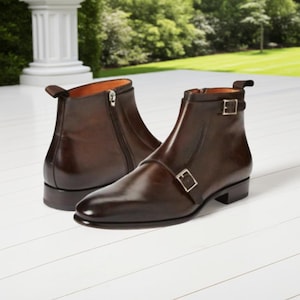 May include: A pair of brown leather ankle boots with a polished finish. One boot has a side zip, the other a buckle closure. The boots are set against a white surface with a blurred green background.