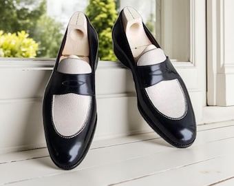 May include: A pair of black and white leather loafers with wooden shoe trees inside. The shoes have a glossy finish and a white leather vamp. The loafers are displayed on a white surface near a window with a green background.
