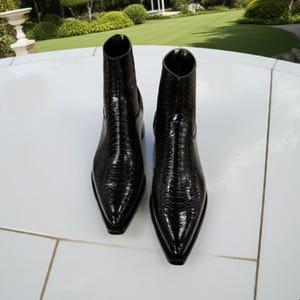 Könnte beinhalten: Ein Paar schwarze Stiefeletten mit einem strukturierten, schlangenartigen Muster. Die Stiefel haben eine spitze Zehenpartie und einen mittelhohen Schaft. Die Stiefel sind aus glänzendem Material und stehen auf einer weißen Oberfläche.