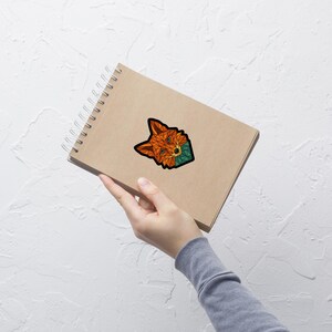 Peut inclure: Un autocollant géométrique représentant une tête de renard avec des accents orange, turquoise et noirs. L'autocollant est collé sur un carnet à spirales avec une couverture beige. Le design du renard est composé de formes angulaires.