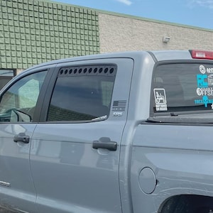 May include: A gray Toyota Tundra truck with black wheels is parked on asphalt. The truck has a TRD 4x4 Off Road logo and stickers on the rear window. The sky is blue with some clouds.