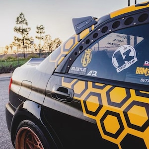 May include: Black and yellow custom car with a honeycomb pattern on the side. The car has several stickers on the window, including "13 Street Team" and "Crawford BAM Shifts". The car is parked on a street.