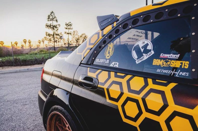 May include: Black and yellow custom car with a honeycomb pattern on the side. The car has several stickers on the window, including "13 Street Team" and "Crawford BAM Shifts". The car is parked on a street.