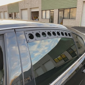 May include: Black car with a custom window vent installed above the rear passenger window. The vent is a black metal plate with multiple circular openings. The car's window reflects the sky and surrounding buildings.