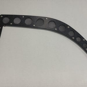 May include: Black metal automotive part with a curved design. It features multiple circular openings with mesh inserts and is secured with small rivets. The part has a right-angle shape on one end and is designed for a vehicle.
