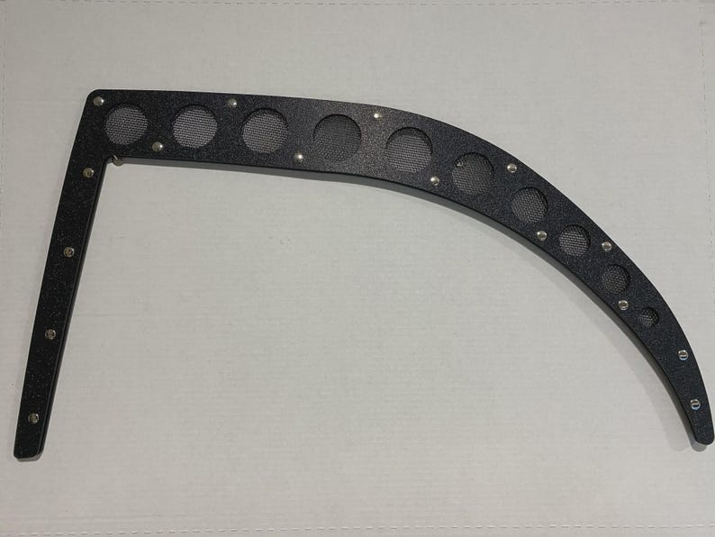May include: Black metal automotive part with a curved design. It features multiple circular openings with mesh inserts and is secured with small rivets. The part has a right-angle shape on one end and is designed for a vehicle.