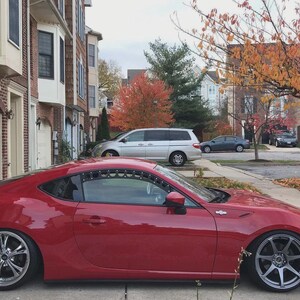 May include: A vibrant red sports car with custom alloy wheels parked on a residential street. The car's low profile and sleek design are prominent. The background features houses, trees with autumn leaves, and other vehicles.