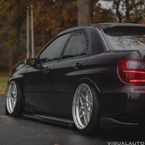 May include: A black sedan with custom silver rims and a lowered suspension. The car has tinted windows and a rear spoiler. The taillights are red and the car is parked on a wet road.