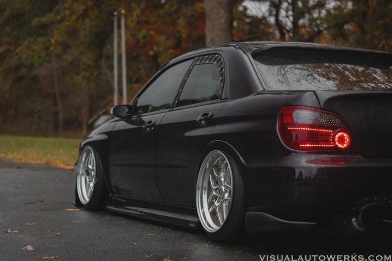 May include: A black sedan with custom silver rims and a lowered suspension. The car has tinted windows and a rear spoiler. The taillights are red and the car is parked on a wet road.