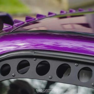 May include: Close-up of a purple car roof with a black trim featuring circular cutouts. The roof has a glossy finish and is adorned with triangular spikes along the edge, adding a unique aesthetic to the vehicle.