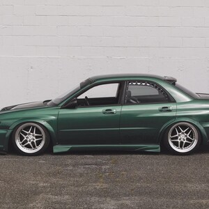May include: A dark green sedan with custom silver alloy wheels and a lowered suspension. The car has a spoiler and tinted windows, parked in front of a white brick wall. The car's body is a deep emerald green.
